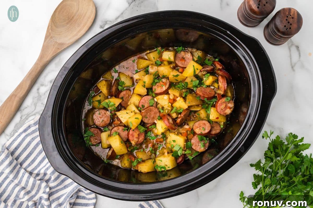 Slow Cooker Sausage and Potatoes 9 a horizontal image of sausages and potatoes in slow cooker in a crock pot with salt and pepper shakers, a wooden spoon and parsley next to it.