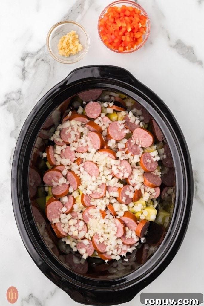 Slow Cooker Sausage and Potatoes 7 a prep image of a crockpot full of cubed potatoes, chopped onion, and sausage with a bowl of red bell pepper and garlic in bowls next to it.