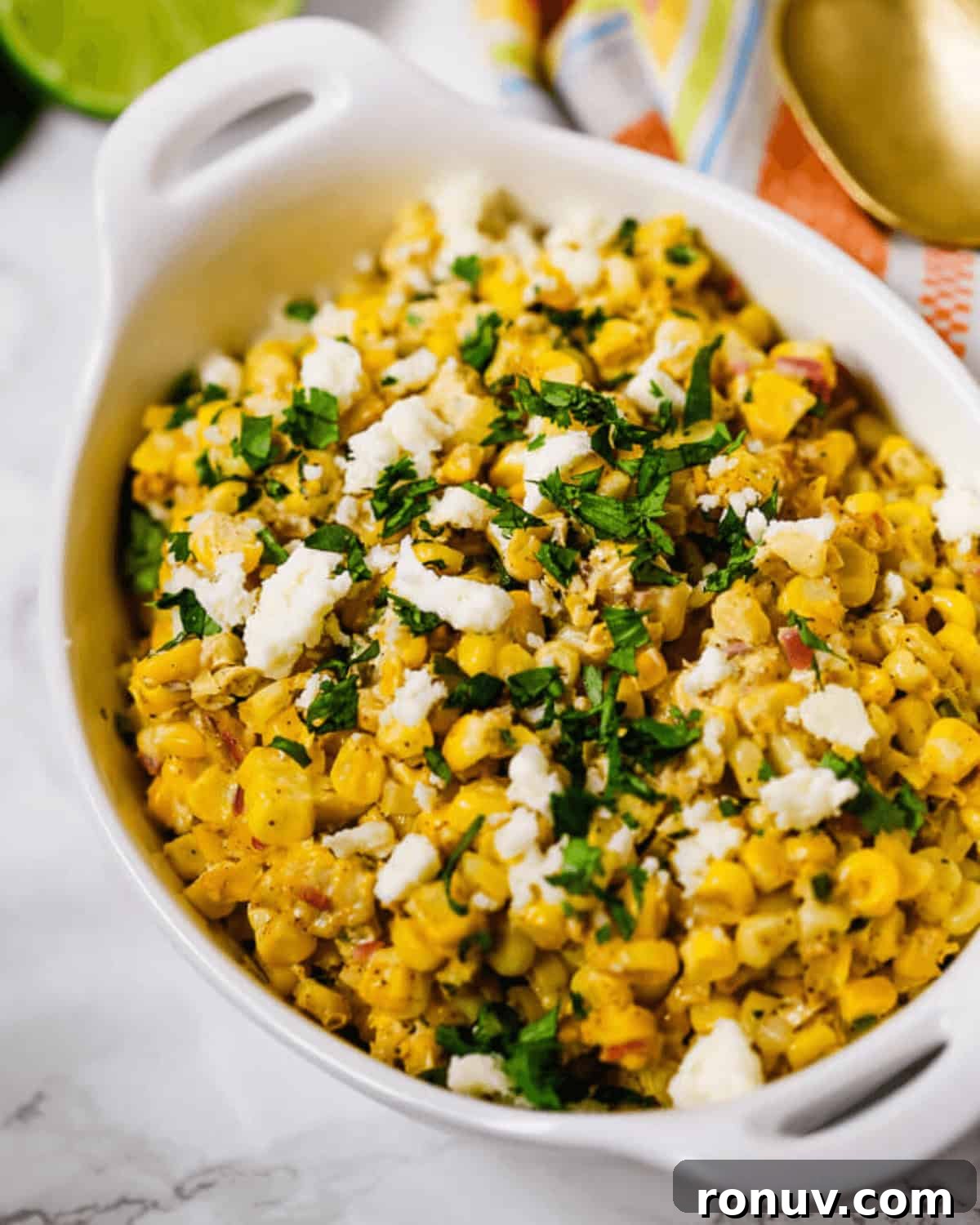 Elote Salad: Mexican Street Corn Recipe 7 A white casserole dish filled with seasoned Elote Salad—corn topped with crumbled cheese and chopped herbs—sits on a marble surface.