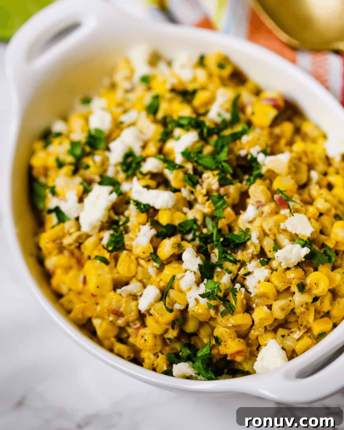 Elote Salad: Mexican Street Corn Recipe 3 A white dish filled with Elote Salad—Mexican street corn topped with crumbled cheese and chopped herbs—rests on a marble surface.