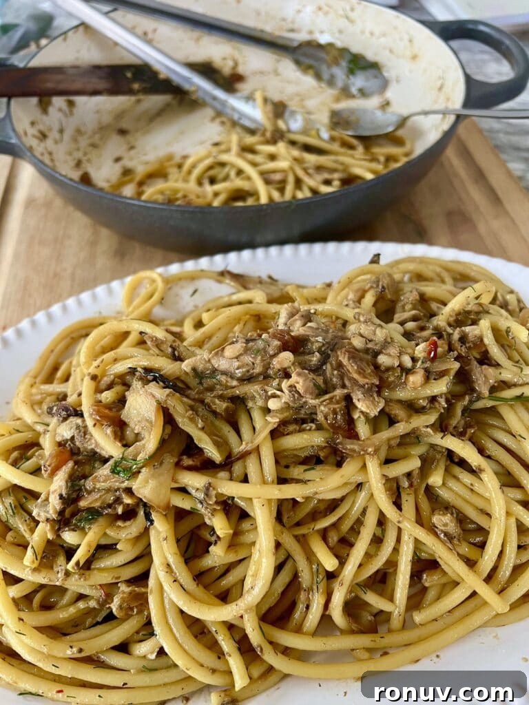 Authentic Sicilian Pasta con Sarde Recipe 4 Pasta con sarde recipe in white platter with pan of it in the background with spoon, tongs and wooden spatula in it.
