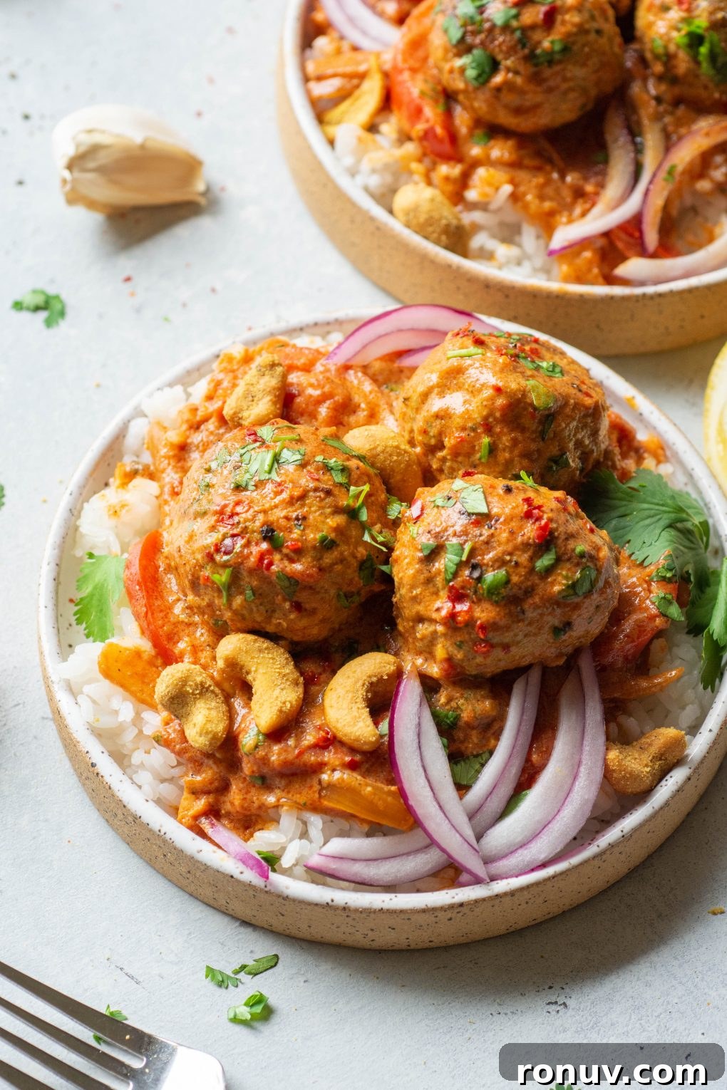 Side angle shot of a small white speckled plate on a light colored background filled with white rice topped with 3 meatballs in a vibrant red curry sauce. Topped with chopped cilantro, thin slices of red onion, and curried cashews.
