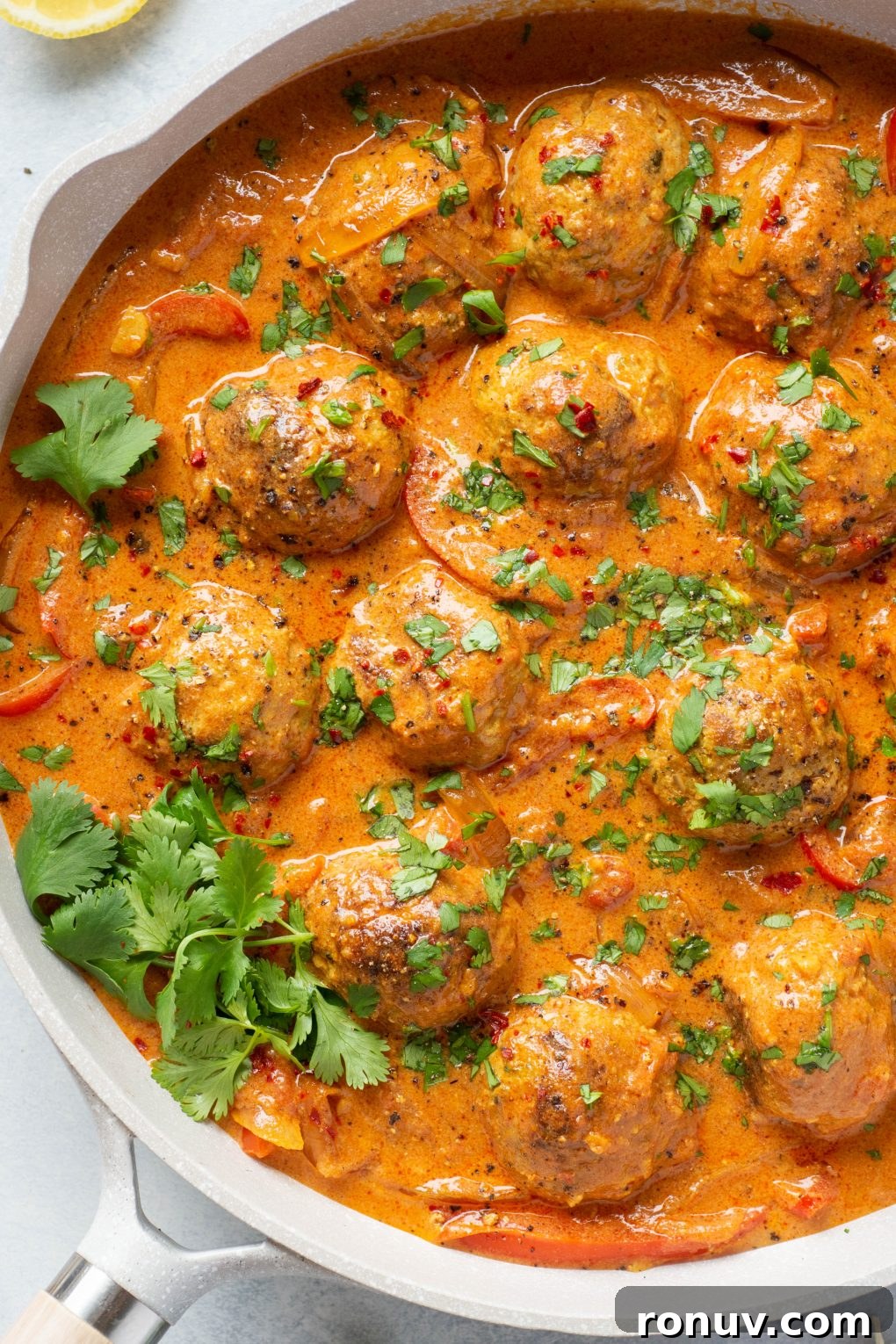 Overhead shot of a large white skillet filled with paleo meatballs in a vibrant red curry sauce. Topped with chopped fresh cilantro on a light colored background.