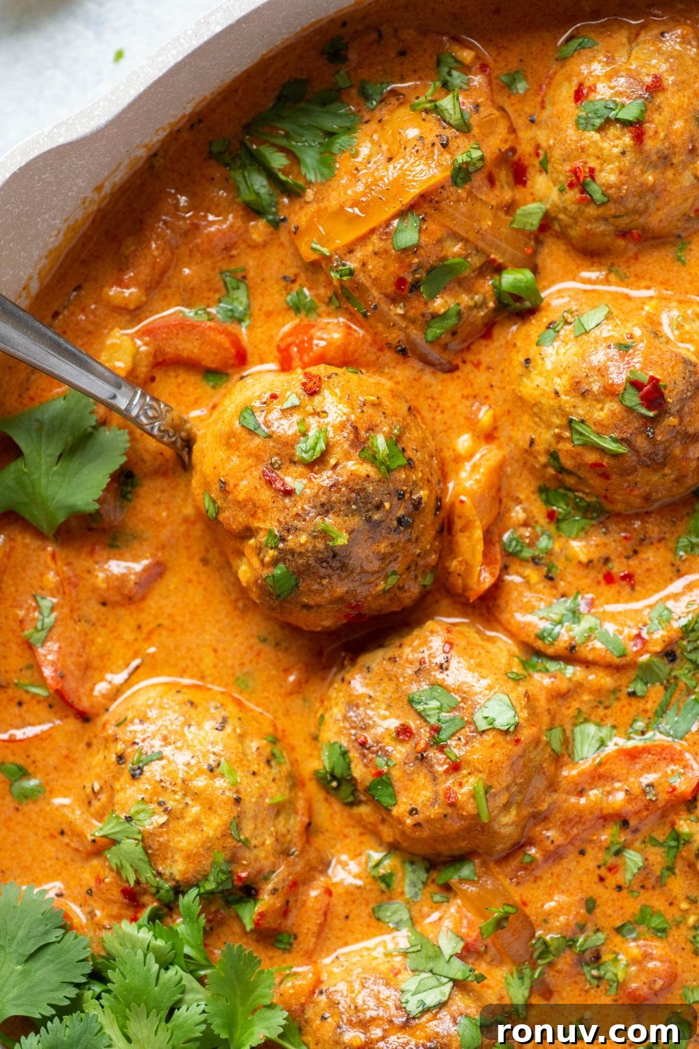 Overhead shot of a spoon holding a single paleo meatball in a skillet of meatballs and vibrant red curry sauce.