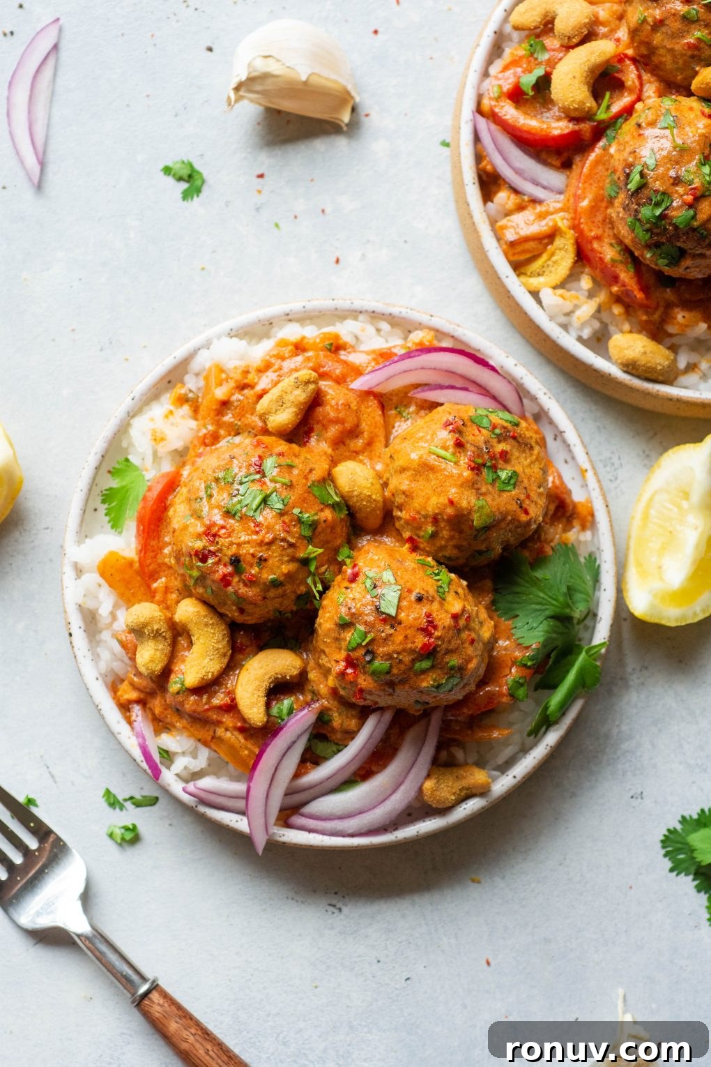Overhead shot of a small white speckled plate filled with white rice and 3 paleo meatballs in vibrant red curry sauce. Topped with chopped cilantro, thinly sliced red onion, and scattered cashews.