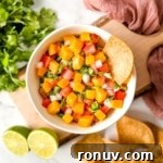 A white bowl of mango pico de gallo surrounded by tortilla chips and limes.