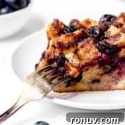 A slice of bread pudding with blueberries on a white plate, with a fork cutting into it.
