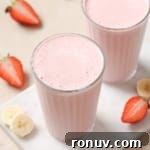 Two glasses of pink strawberry banana smoothie with sliced strawberries and bananas around.
