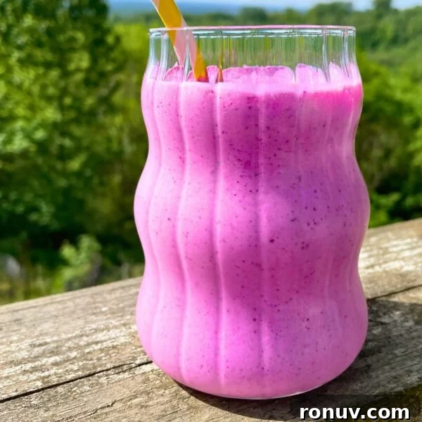 Blueberry pineapple smoothie in a glass with a yellow straw on a wood railing outside with trees behind it.