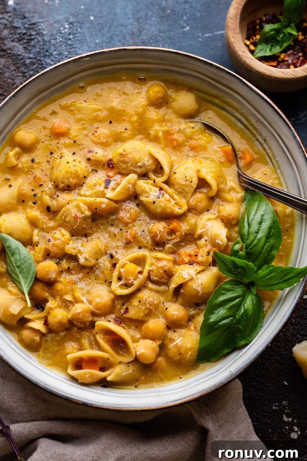 Pasta e Ceci Recipe: Classic Italian Pasta with Chickpeas 7 A bowl filled with pasta e ceci soup and fresh herbs on top. The bowl is sitting on a dark background with a napkin and small bowl of red pepper flakes arranged around it.