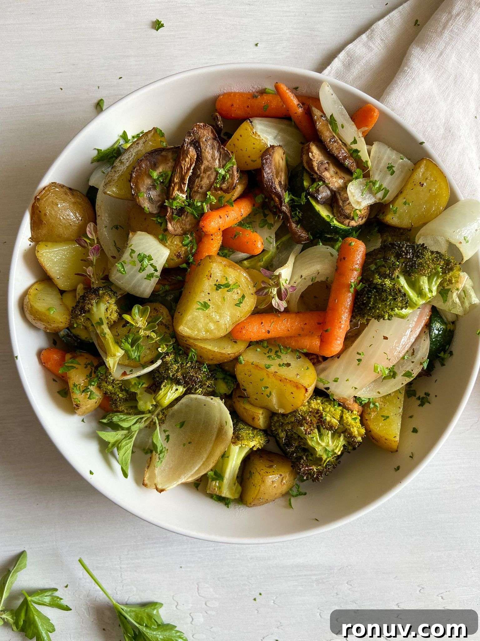 Crispy Oven-Roasted Vegetables with Garlic and Herbs 2 A white bowl filled with oven roasted vegetables including potatoes, carrots, broccoli, onions, and mushrooms, garnished with chopped herbs on a light surface.