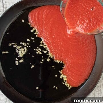 Ricotta-Stuffed Pasta Shells Recipe 8 Tomato puree being poured into a black skillet containing oil and garlic.