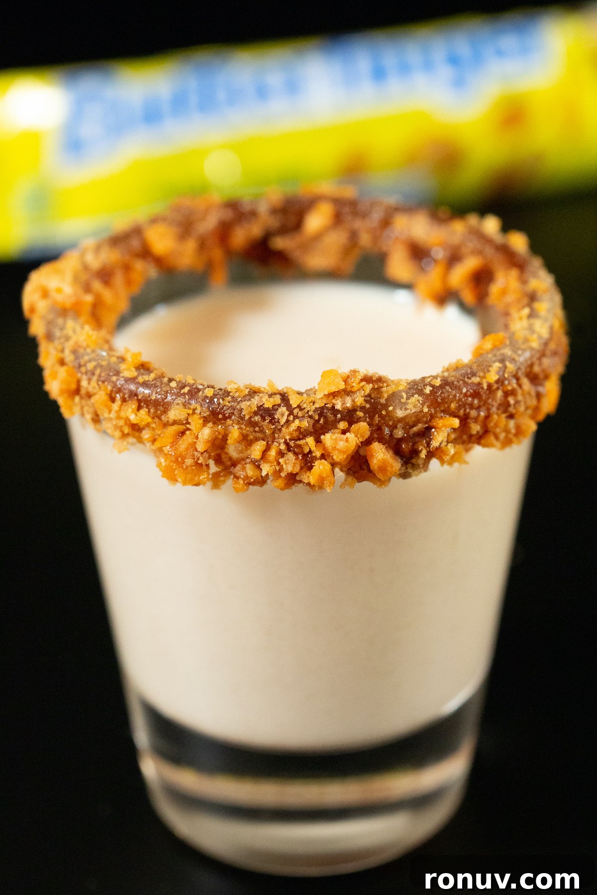 Butterfinger Shot Recipe: Candy Bar Cocktail 2 A shot glass is filled with a light brown Butterfinger shot. The rim is coated in chocolate sauce and crushed candy bar.