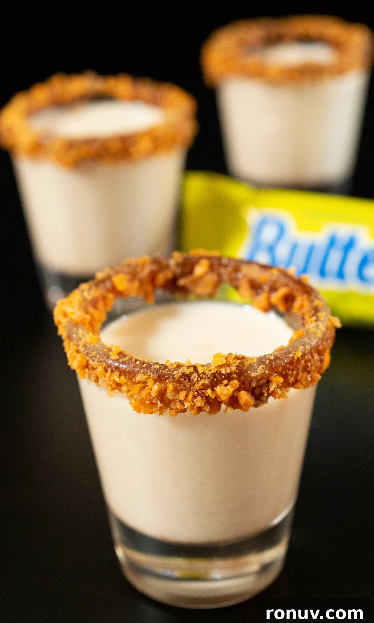 Butterfinger Shot Recipe: Candy Bar Cocktail 4 Three shot glasses all filled with light brown Butterfinger shots. Their rims are coated in chocolate sauce and crushed pieces of the candy bar.