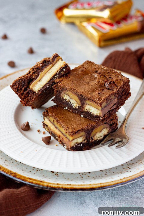 Homemade Fudgy Twix Brownies Recipe 2 3 Twix brownies and a fork stacked on a white ribbed plate over a mottled grey backdrop. There are 2 Twix bars, a brown linen napkin and chocolate chips scattered in the background.