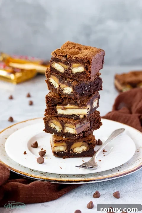 Homemade Fudgy Twix Brownies Recipe 9 A stack of Twix brownies on a white ribbed plate with chocolate chips nearby.