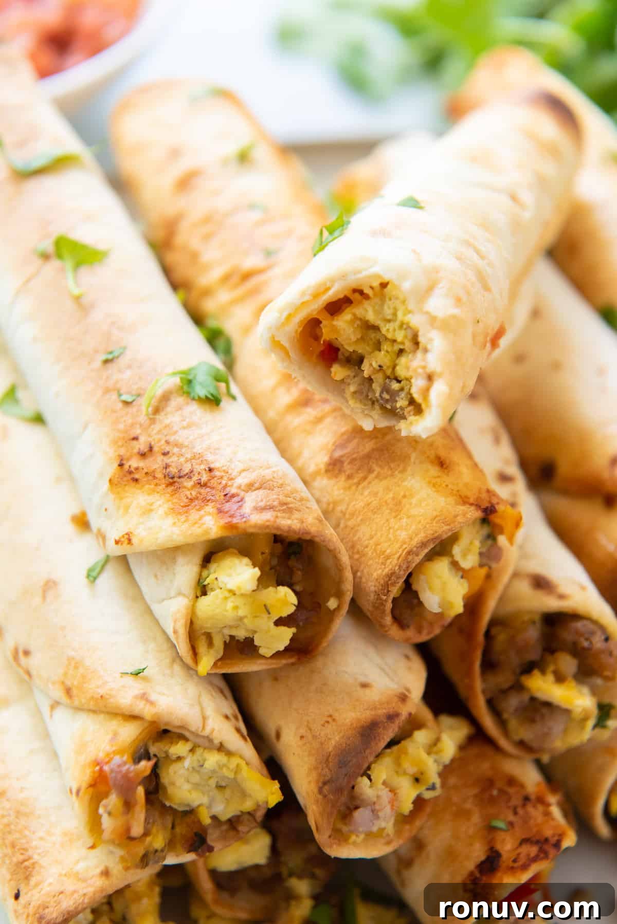 Crispy Oven-Baked Breakfast Taquitos Recipe 8 An egg and sausage breakfast taquito with a bite missing stacked on top of others.