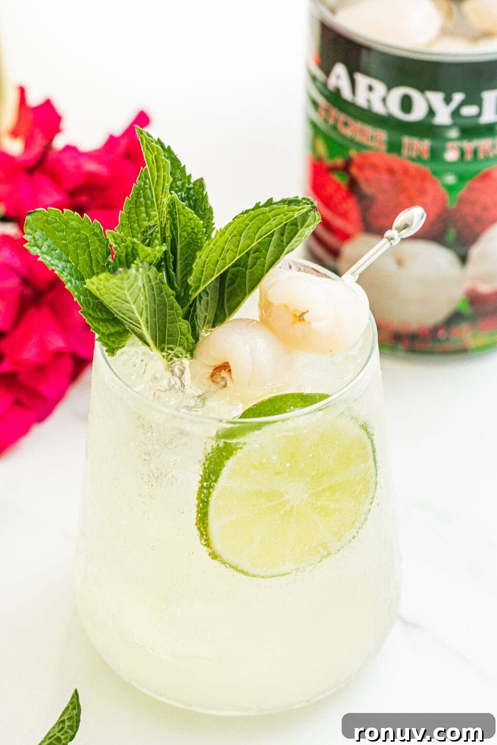 a stemless wine glass filled with lychee cocktail spritz, garnished with two lychees, a lime wheel, fresh mint, can of lychees, purple plant in the background 