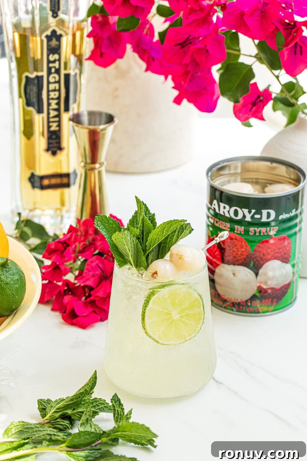 a stemless wine glass filled with lychee cocktail spritz, garnished with two lychees, a lime wheel, fresh mint, can of lychees, purple plant in the background, bottle of elder flower liqueur and a gold jigger