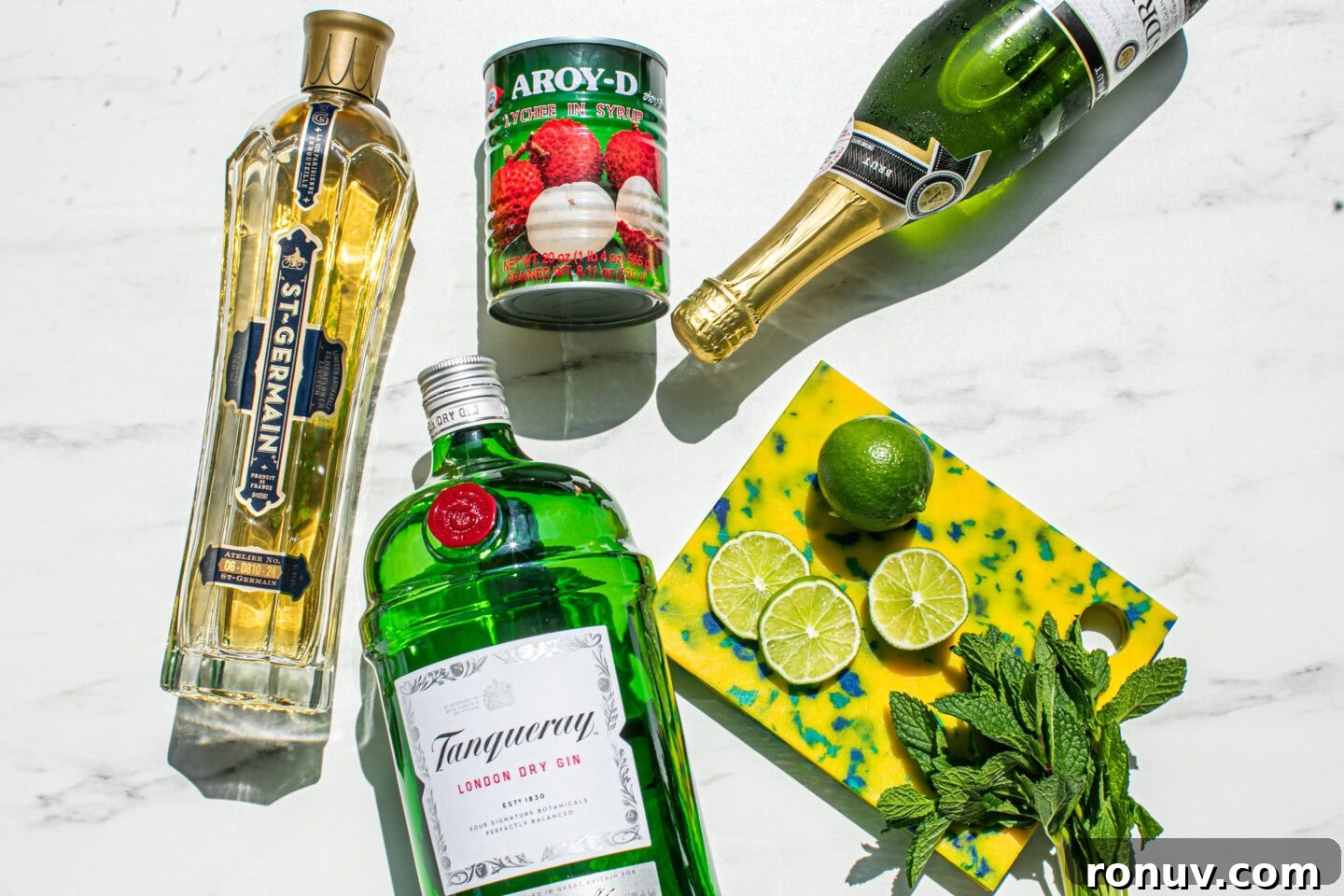 bottle of st germain elderflower liqueur, gin bottle, can of lychees, lime wheels, mint sprigs and a bottle of prosecco 
