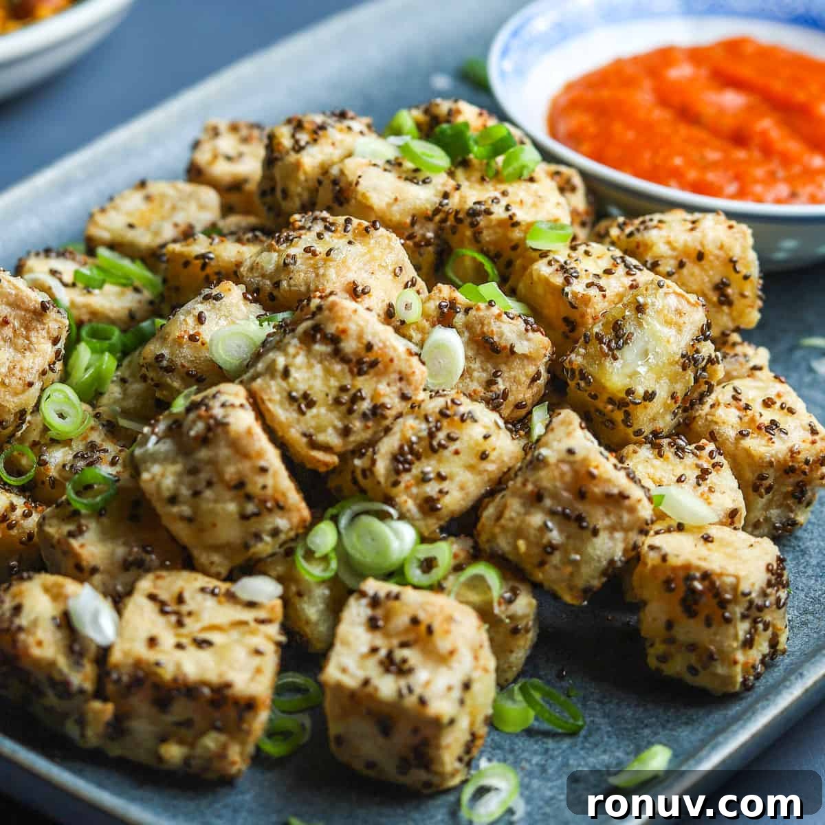 Crispy Air Fryer Tofu Recipe with Chia Seeds 2 photo of a blue oblong plate filled with crispy air fried tofu with spring onion sprinkled on top and a bowl of kimchi sauce in the background.