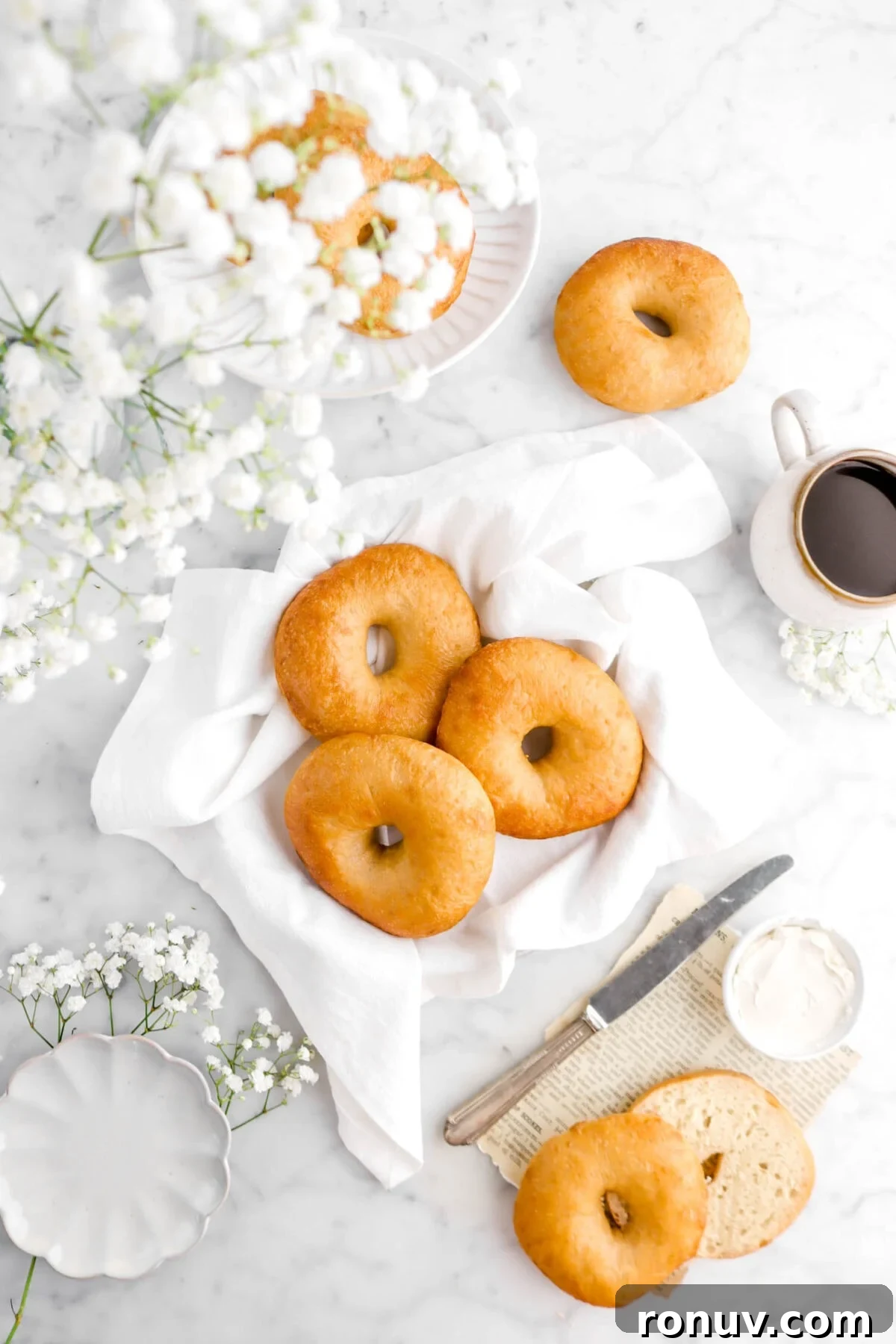 How to Make Sourdough Bagels at Home 2 bagels on white napkin with more bagels around, one cut open beside, with a knife and cream cheese beside, a cup of coffee, and flowers around