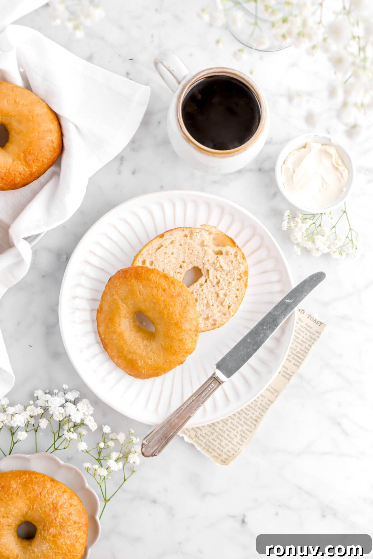 How to Make Sourdough Bagels at Home 3 overhead shot of cut open bagel on white plate with knife, bowl of cream cheese, flowers, and cup of coffee
