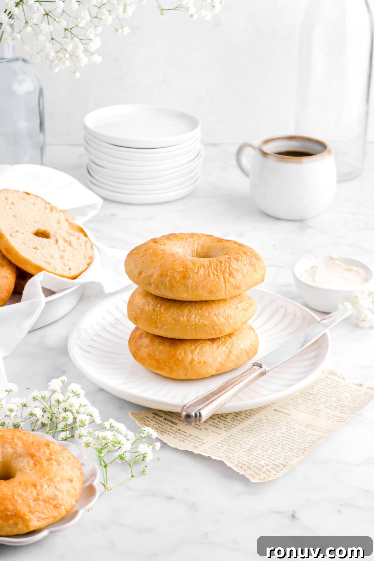 How to Make Sourdough Bagels at Home 4 three stacked bagels on white plate with cup of coffee, stack of white plates, and flowers