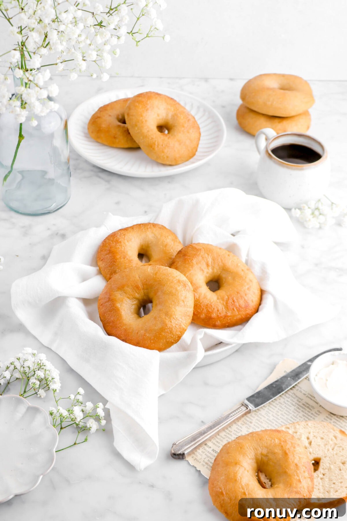 How to Make Sourdough Bagels at Home 5 angled shot of three bagels in pie plate with white napkin, four bagels behind with a glass vase of flowers, and cup of coffee