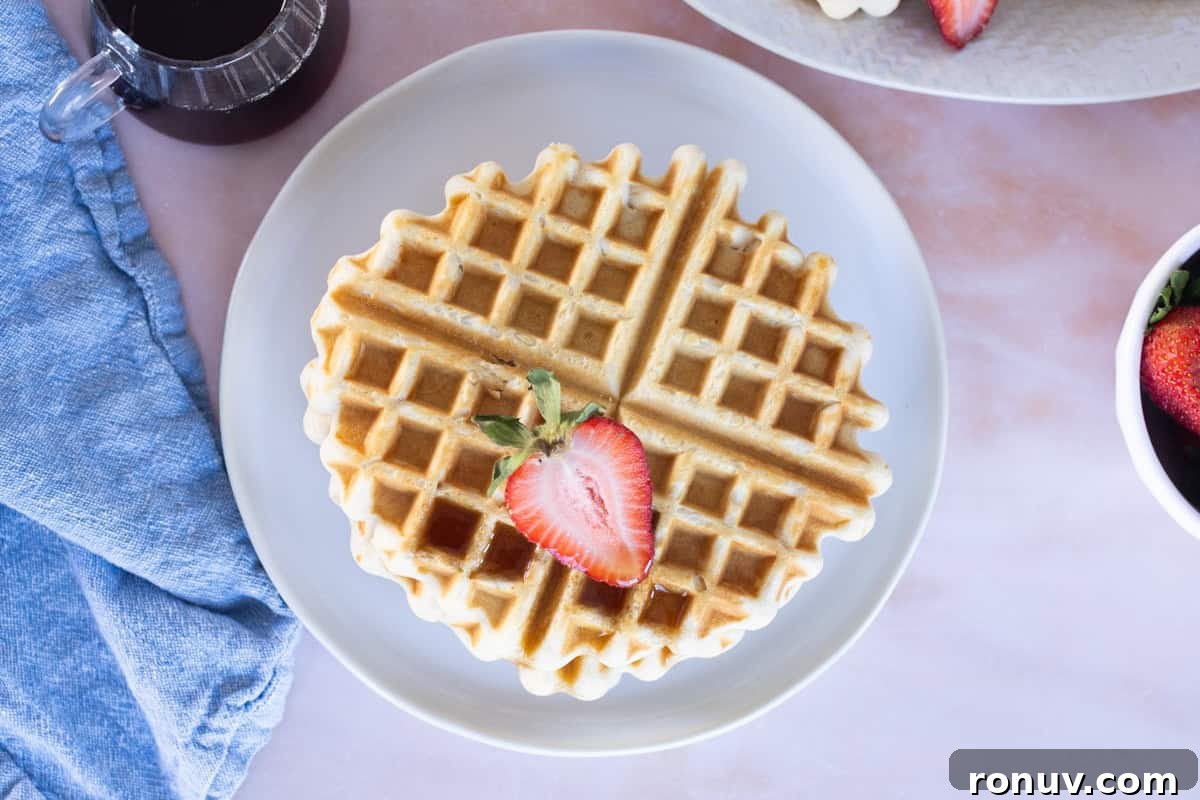 Fluffy Homemade Waffles Recipe 10 A round Easy Waffle on a white plate topped with sliced strawberry, served with syrup, a blue napkin, and a bowl of strawberries nearby.