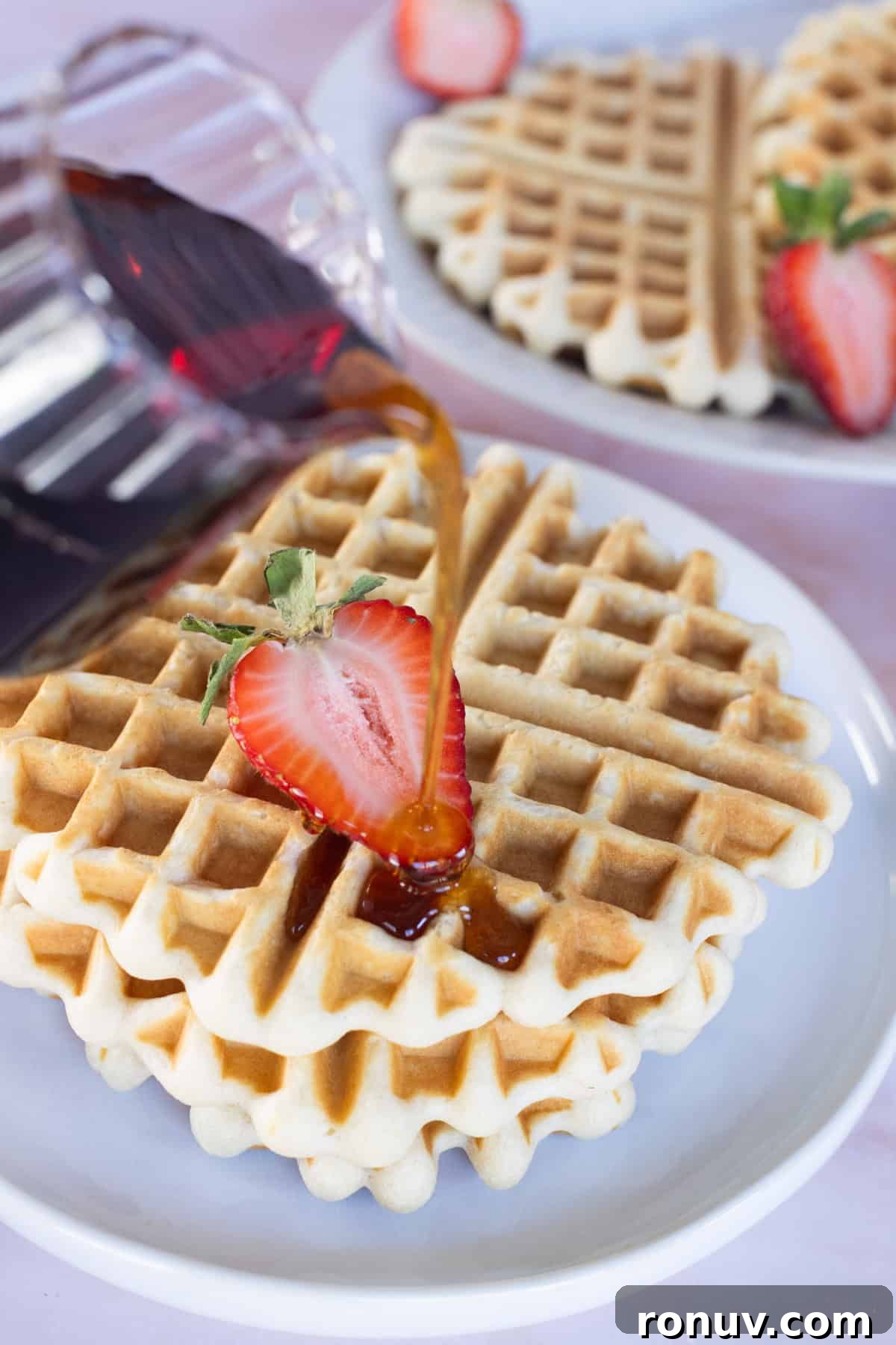 Fluffy Homemade Waffles Recipe 4 Syrup is being poured onto a stack of plated waffles topped with a sliced strawberry. Another plate of waffles sits in the background.