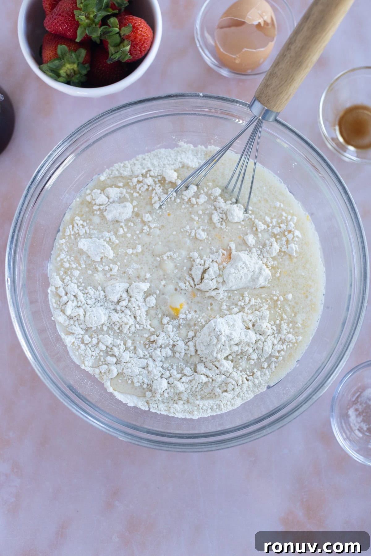 Fluffy Homemade Waffles Recipe 7 A mixing bowl with flour, milk, and egg being whisked for easy waffles; nearby are a bowl of strawberries, an eggshell, vanilla extract, and an empty glass bowl.