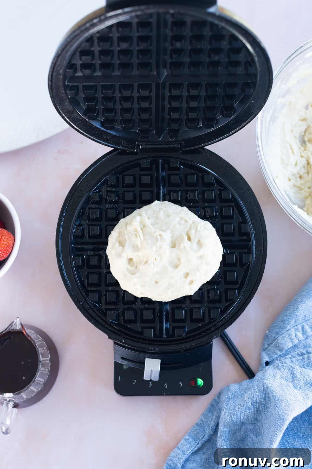 Fluffy Homemade Waffles Recipe 8 A portion of easy waffle batter sits on an open waffle maker, surrounded by a bowl of batter, strawberries, syrup, and a blue cloth.