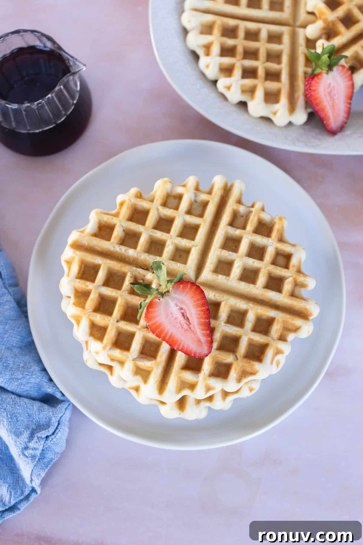 Fluffy Homemade Waffles Recipe 9 A plate with an Easy Waffles round waffle topped with a halved strawberry, next to a pitcher of syrup and a partial plate of waffles in the background.