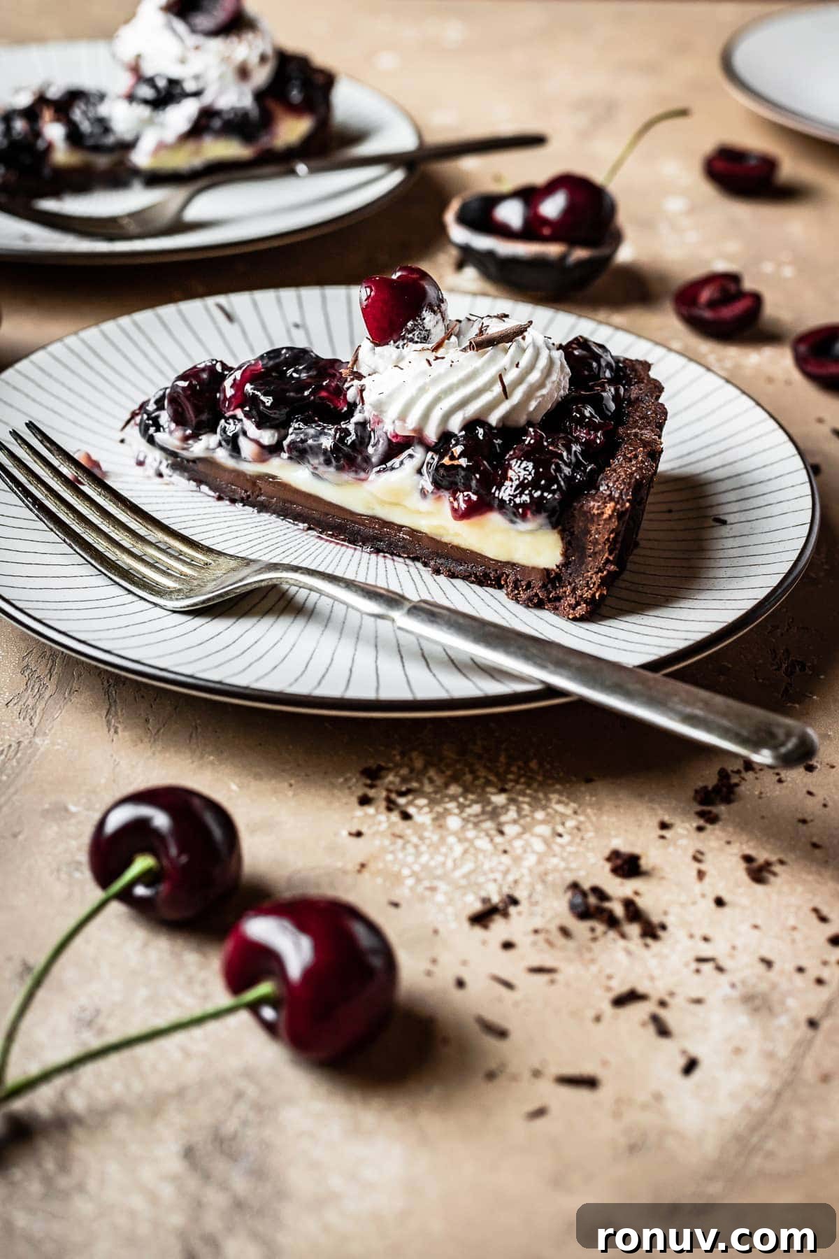 Decadent Chocolate Cherry Tart Recipe 4 A side view of a slice of tart on a plate. Another plate is in the background, and cherries are scattered around the brown backdrop.