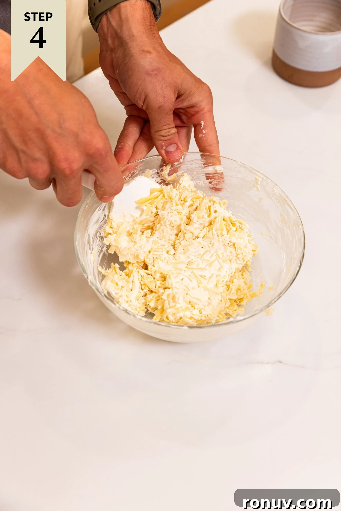 Fresh Tomato Galette Recipe with Basil 11 Hand holding spatula mixing together cream cheese and shredded cheese in small glass bowl on white surface.